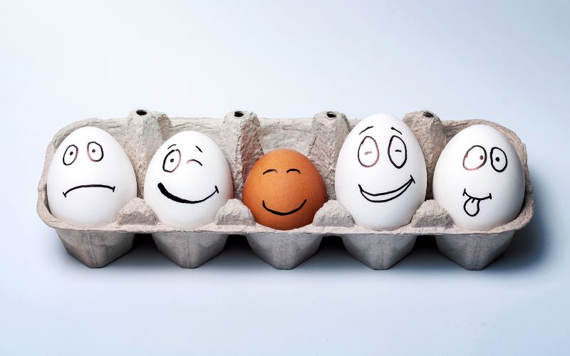 Fünf mit Smileys bemalte Eier in einem Eierkarton, das mittlere Ei ist braun und hat ein lachendes Gesicht.