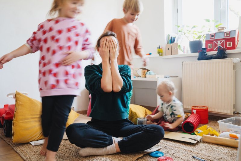 Mutter auf dem Boden sitzend, hält sich das Gesicht zu, drei Kinder rennen um sie herum mit Baby liegend auf dem Sofa